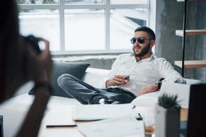 traumhafte Atmosphäre. Foto auf der linken Seite. junger kurzhaariger Mann mit Sonnenbrille sitzt auf der Couch im Büro