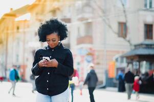 attraktiv glücklich dunkelhäutig Modell- mit afro Frisur und Nasenring, posieren draußen gegen städtisch Hintergrund während ihr Morgen gehen, suchen Nieder mit schüchtern Lächeln zeigen ihr Weiß Zähne. Fackel Sonne foto