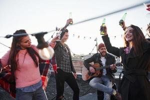 Flaschen auf und singen. Gruppe junger Leute, die auf einem Dach mit etwas Alkohol und Gitarrenspiel feiern? foto
