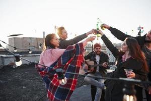 erhebe dich und klopfe auf die Brille. Gruppe junger Leute, die auf einem Dach mit etwas Alkohol und Gitarrenspiel feiern? foto