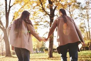 Blick von der Rückseite junger brünette Zwillingsmädchen, die Händchen halten und im lässigen Mantel im sonnigen Herbstpark auf verschwommenem Hintergrund spazieren gehen