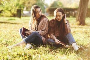 Junge brünette Zwillingsschwestern sitzen dicht beieinander mit geschlossenen Augen auf dem Gras, Beine leicht in die Knie gebeugt und gekreuzt, halten braune Bücher und tragen einen lässigen Mantel im Herbstpark im Hintergrund foto