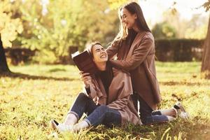 lachende brünette Zwillingsmädchen, die auf dem Gras sitzen. einer von ihnen hält ein braunes Buch, der andere lehnt sich im herbstlichen sonnigen Park auf verschwommenem Hintergrund mit den Händen von hinten auf die Schultern der Schwestern foto