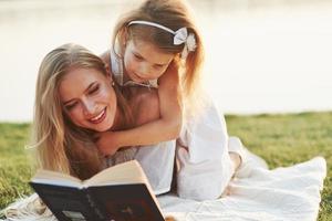 es erregt eine ganz besondere Aufmerksamkeit. Mutter und Tochter, die an einem sonnigen Tag ein Buch lesen, das auf dem Gras mit See im Hintergrund liegt foto