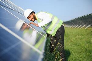 Porträt von jung indisch Mann Techniker tragen Weiß schwer Hut Stehen in der Nähe von Solar- Paneele gegen Blau Himmel.Industrie Arbeiter Solar- System Installation, verlängerbar Grün Energie Generation Konzept. foto