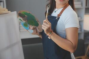 abgeschnitten Bild von weiblich Künstler Stehen im Vorderseite von ein Staffelei und Eintauchen Bürste in Farbe Palette foto