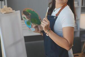 abgeschnitten Bild von weiblich Künstler Stehen im Vorderseite von ein Staffelei und Eintauchen Bürste in Farbe Palette foto