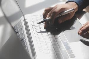 abgeschnitten Bild von ein jung Mann Arbeiten auf seine Laptop im ein Kaffee Geschäft, Rückseite Aussicht von Geschäft Mann Hände beschäftigt mit Laptop beim Büro Schreibtisch, jung männlich Schüler Tippen auf Computer Sitzung beim hölzern Tabelle foto