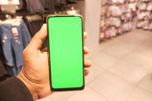 halten Clever Telefon mit Grün Bildschirm gegen Einkaufen Einkaufszentrum Hintergrund foto