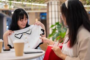 zwei asiatisch Frauen sitzen im ein Cafe beim ein Einkaufszentrum, einer aufgeregt zeigen aus ein Neu Hemd zu ihr Freund. foto