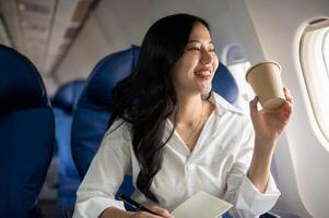 ein elegant asiatisch Geschäftsfrau ist nippen Kaffee während suchen beim das Aussicht draußen das Flugzeug Fenster. foto