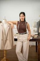 ein zuversichtlich asiatisch weiblich Mode Designer ist Stehen im ihr Atelier Studio mit ein Schneider Attrappe. foto