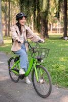 ein glücklich jung asiatisch weiblich Hochschule Schüler ist genießen Reiten ihr Fahrrad im ein Grün Park. foto