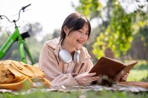 ein glücklich asiatisch Frau ist genießen lesen ein Buch auf ein Picknick Tuch, während chillen im ein Grün Park. foto