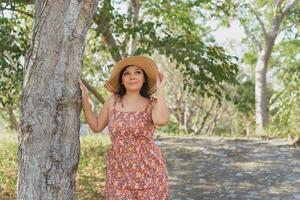 Frau tragen Hut und Kleid mit Blumen bewundern Natur im ein Öffentlichkeit Park schließen zu ein Baum. sonnig Sommer- Tag. foto