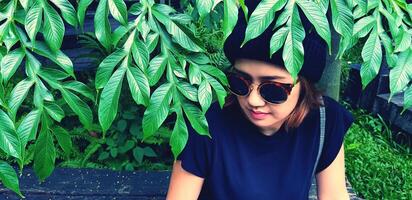 asiatisch Frau tragen Sonnenbrille und wolle Hut Sitzung auf hölzern Bank unter Baum mit Grün Pflanze Hintergrund. Mädchen im beiläufig Kleid Stil im Jahrgang Ton Farbe mit Kopieren Raum. foto