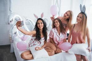 Luftballons und Hasenohren. Junggesellinnenparty. Vier Mädchen in rosa und weißer Kleidung sitzen auf dem Sofa und machen Selfie