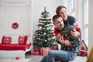 Fröhliche junge Leute sitzen mit Weihnachtsdekoration auf der Fensterbank im Zimmer und umarmen sich foto