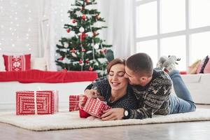 Kerl gibt seiner Frau Weihnachtsgeschenk. schönes junges Paar, das auf dem Wohnzimmer mit grünem Feiertagsbaum im Hintergrund liegt foto