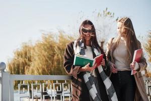 Zwei Studentinnen gehen auf der Brücke im Park spazieren, während sie Notizblöcke halten foto