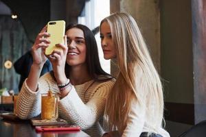 im Restaurant. Zwei Freundinnen sitzen mit gelbem Getränk drinnen und benutzen das Smartphone foto
