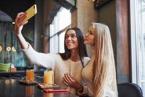 aufrichtige Brünette hat ein schönes Lächeln. Junge Freundinnen machen Selfie im Restaurant mit zwei gelben Getränken auf dem Tisch foto