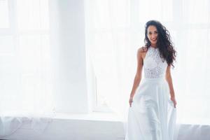 Luxus tragen. Schöne Frau im weißen Kleid steht im weißen Raum mit Tageslicht durch die Fenster