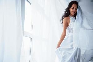 elegantes Tragen. Schöne Frau im weißen Kleid steht im weißen Raum mit Tageslicht durch die Fenster