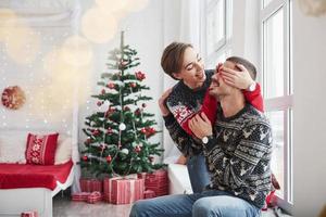 Rate mal, was ich dir schenken werde. Fröhliche junge Leute sitzen auf der Fensterbank im Zimmer mit Weihnachtsschmuck foto