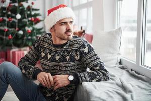 auf der Fensterbank gelehnt. Foto des Mannes in Weihnachtsmütze und Urlaubskleidung sieht durch das Fenster. Weihnachtsbaum im Hintergrund