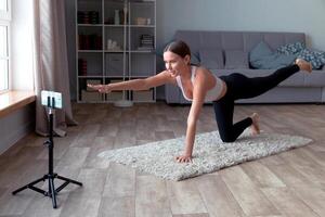 Frau im schwarz Gamaschen und ein Weiß oben tut Yoga beim Zuhause auf das Teppich, Aufzeichnung ein online Fitness zum ihr Publikum mit ein Smartphone Stehen auf ein Stativ foto