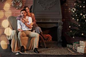 Luxusportrait. Schönes junges Paar auf dem Stuhl, der sich in einem Luxuszimmer umarmt, das mit Weihnachtsbaum und Girlanden dekoriert ist foto