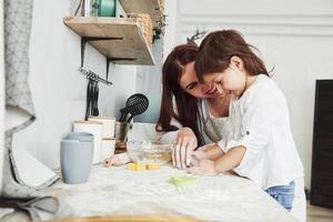 schöne Frau lächelt. glückliche tochter und mutter bereiten zusammen backwaren zu. kleiner Helfer in der Küche foto