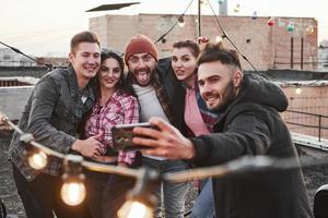 Kerl in der Mitte macht lustiges Gesicht. Gruppe junger fröhlicher Freunde, die Spaß haben, sich umarmen und Selfies auf dem Dach mit dekorativen Glühbirnen machen foto