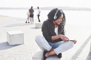 Porträt eines schönen jungen hübschen afroamerikanischen Mädchens, das am Strand oder See sitzt und Musik in ihren Kopfhörern hört