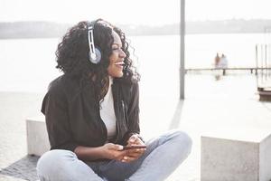 Porträt eines schönen jungen hübschen afroamerikanischen Mädchens, das am Strand oder See sitzt und Musik in ihren Kopfhörern hört