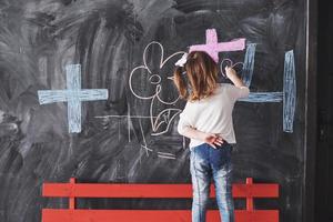 lockiges kleines Mädchen, das mit Kreidefarbe an der Wand zeichnet. Werke des Kindes. süße Schülerin schreibt auf Tafel foto
