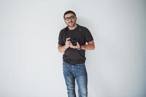 Ein junger Hipster-Mann in Okularen hält eine DSLR-Kamera in den Händen, die vor einem weißen Wandhintergrund steht foto