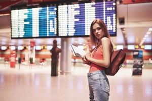 Schönes junges Touristenmädchen mit Rucksack im internationalen Flughafen, in der Nähe der Fluginformationstafel