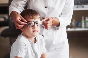 Süßes Kind schaut in die Kamera, während die Ärztin ihm eine neue Brille trägt