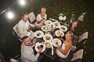 Alle schauen bitte in die Kamera. Ansicht von oben. Gruppe von Freunden in der eleganten Kleidung haben ein luxuriöses Abendessen foto