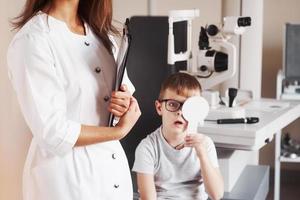 er ist schockiert, dass er diese Worte lesen kann. Frau mit Dokumenten testet die Sehschärfe eines kleinen Jungen foto