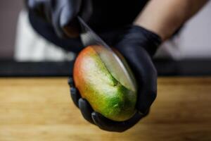 Person mit schwarz Handschuhe Schneiden und Schneiden Mango zum Smoothie Holz Schneiden foto