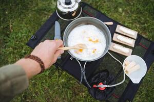 ein Kerl Braten Eier im ein braten schwenken beim ein Campingplatz, Kochen auf ein Wanderung Morgen Frühstück im Natur, rühren mit ein Gabel Glasur, Tourist Utensilien Aussicht von über. foto