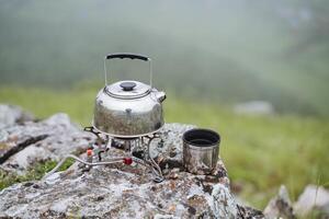 Tourist Kessel steht auf das Brenner Sieden Wasser, brauen Tee im Natur auf ein Wanderung, Metall Camping Utensilien, Becher steht auf ein Stein, Gas Brenner foto