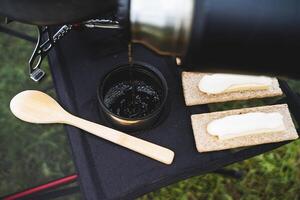 gießen heiß Tee von ein Thermosflasche in ein Glas, ein Aussicht von das Gießen Wasser auf oben von ein Becher, ein hölzern Löffel auf das Tisch, Butter Sandwiches, Essen auf ein Wanderung. foto