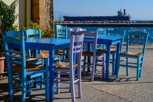 selektiv Fokus auf das schließen Blau hölzern Tabelle und Stuhl beim typisch Fisch Taverne beim Angeln Hafen. groß Schiff auf Hintergrund. ägäisch Insel Chios im Griechenland auf ein Herbst Tag. griechisch Ferien und Ziele foto