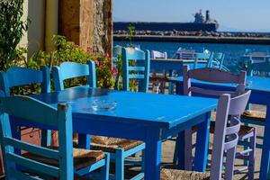 Nahansicht auf hölzern Tabelle und Stuhl im typisch Fisch Taverne beim Angeln Hafen. selektiv Fokus. groß Schiff auf Hintergrund. ägäisch Insel Chios im Griechenland auf ein Herbst Tag. griechisch Ferien und Ziele foto