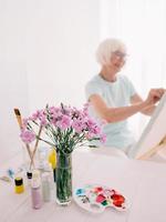 Senior fröhliche Künstlerin in Gläsern mit grauen Haaren, die Blumen in der Vase malen. Kreativität, Kunst, Hobby, Berufskonzept foto