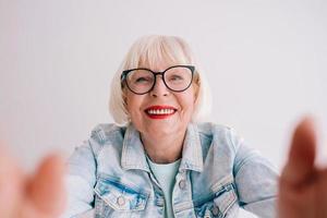 Senior stylische Frau mit grauem Haar und in Brille und Jeansjacke umarmt dich oder macht Selfie. Liebe, Umarmungen, Anti-Age, positives Vibes-Konzept